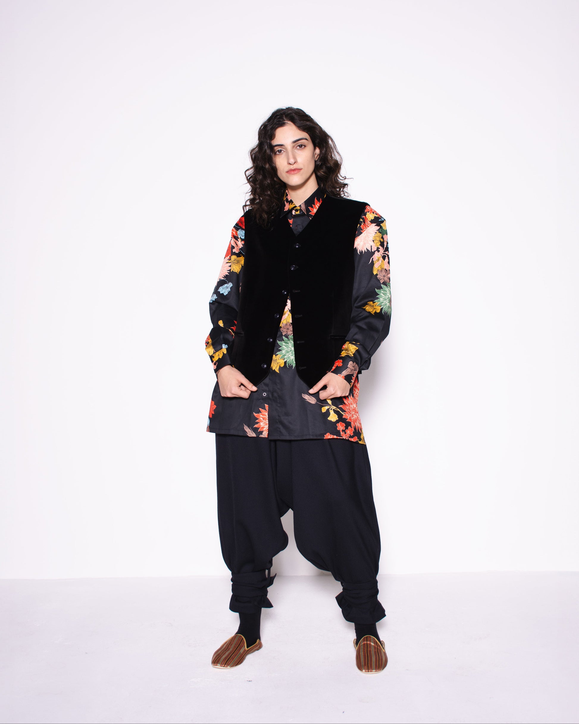 Person wearing a floral shirt, black vest, and dark pants on a white background